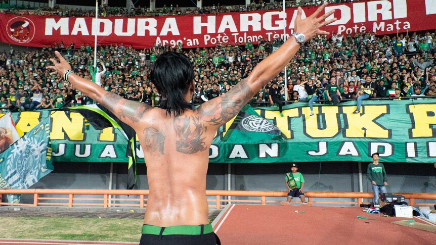 An tattooed entertainer raises his arms and faces the stadium crowd.