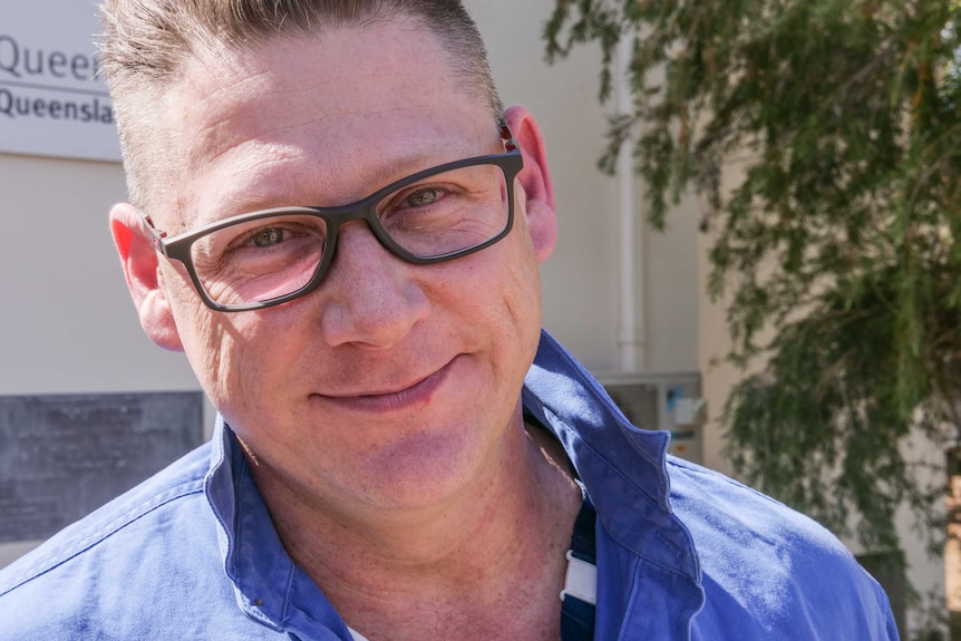 A man with glasses smiles in front of a demountable building.