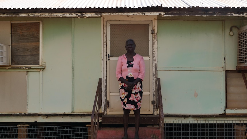 An Indigenous woman in a pink jacket stands in front of a turquoise building with a tin roof