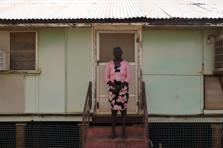 An Indigenous woman in a pink jacket stands in front of a turquoise building with a tin roof