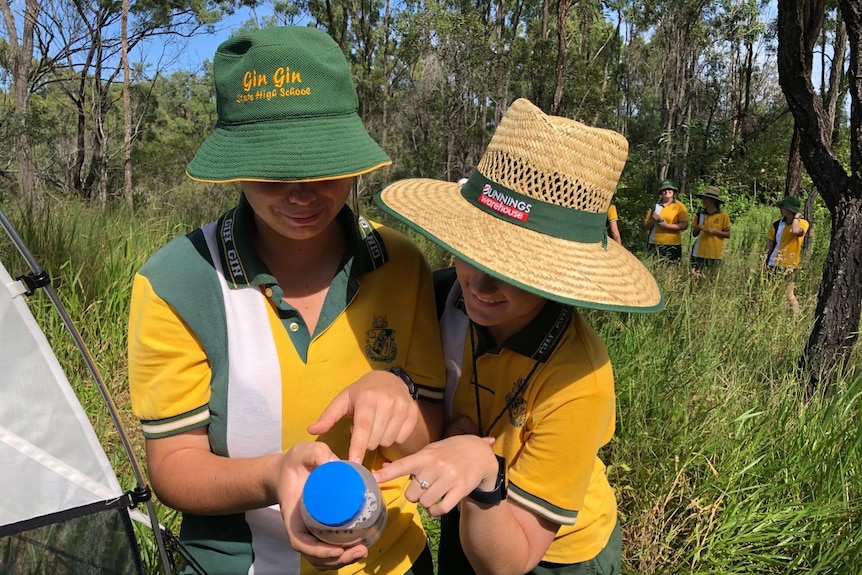 Two school girls wearing hats look down at a bottle in their hands