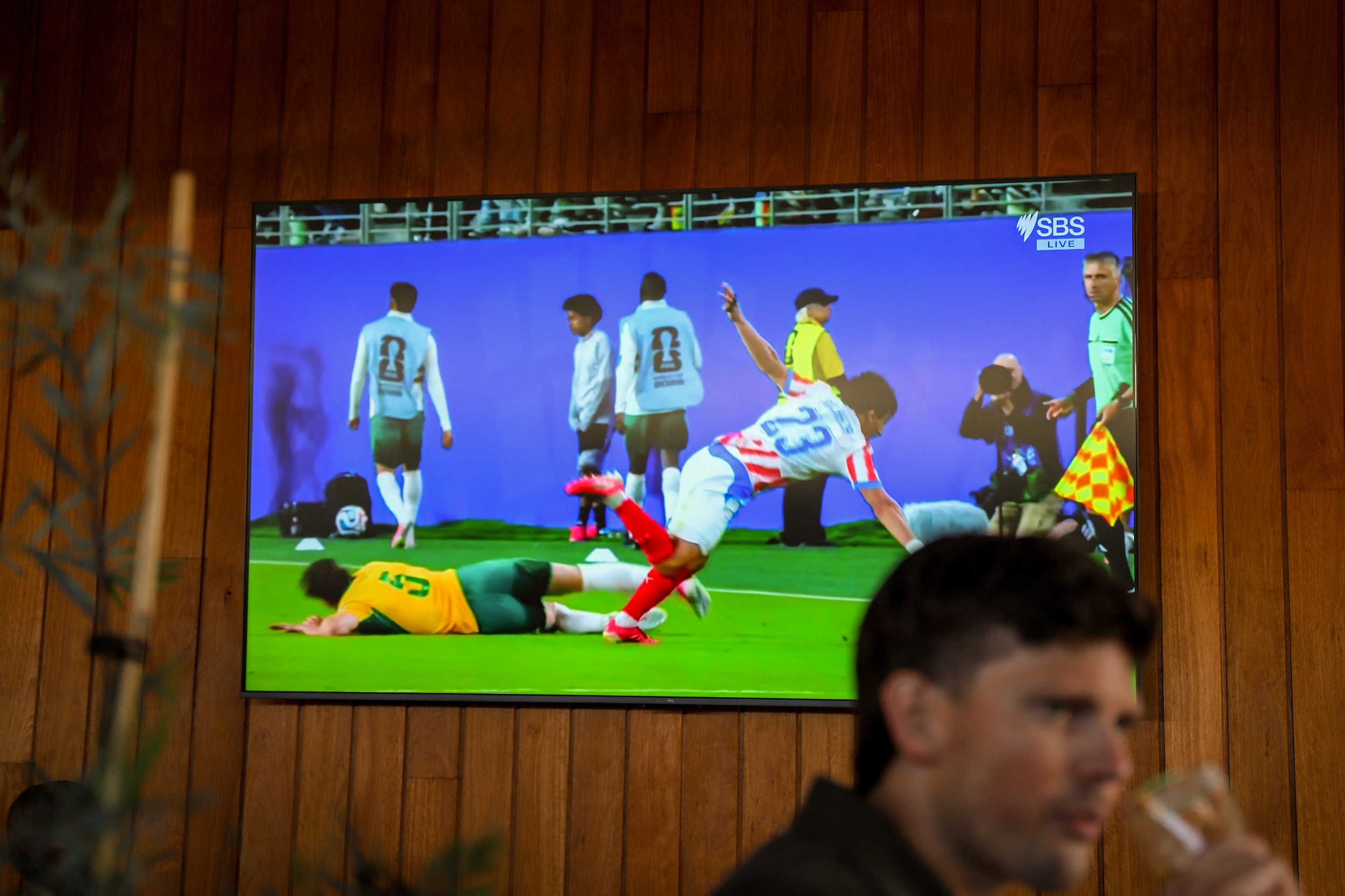 A television plasma screen screening world cup soccer affixed to a wooden pub wall with man drinking beer in foreground