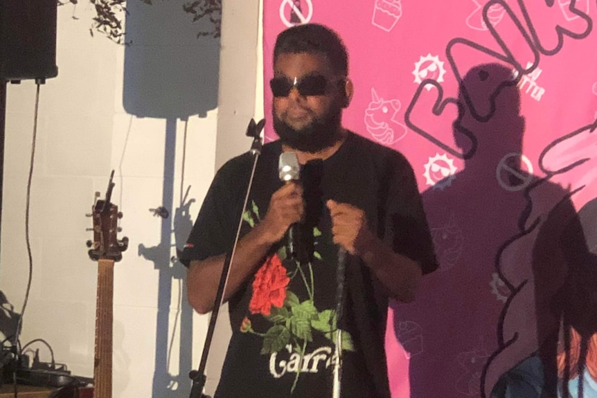 Indigenous man in dark sunglasses, standing in from of pink backboard with microphone