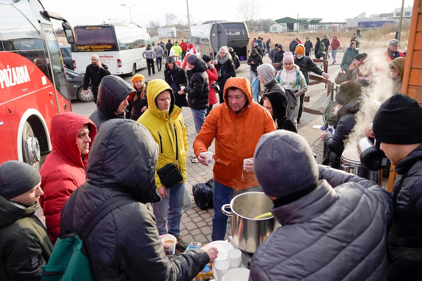 a group of refugees gathers around a man in an orange coat offering coffee as buses and other refugees can be seen behind