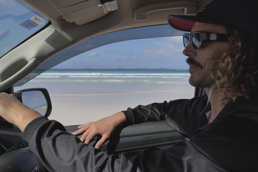 He has curly hair and wear a hat and glasses as he drives along a beach