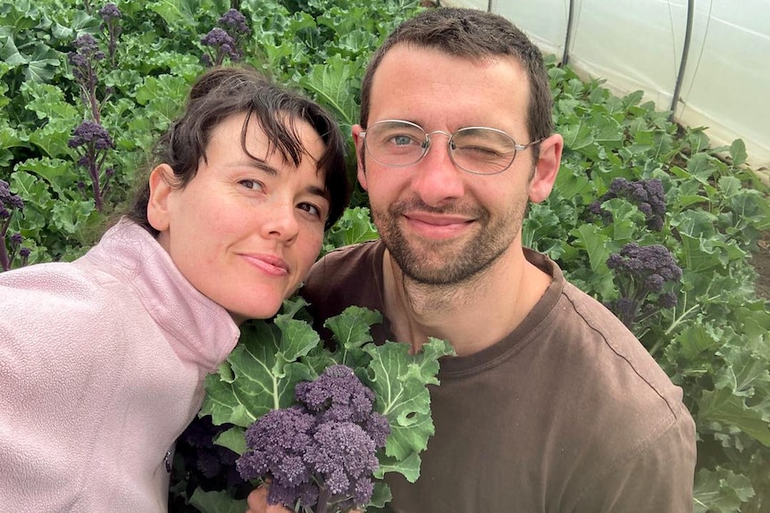 A young smiling couple take a selfie in front of a crop of purple broccoli