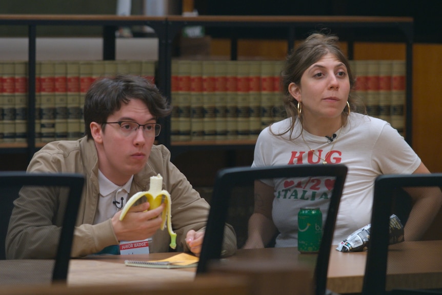 A man in his 20s with glasses eats a banana and a woman in her early 30s in a white tshirt sit at a meeting table, books in back