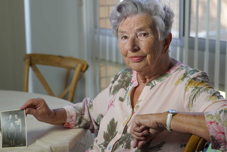 A portrait of a senior woman with grey hair holding a black and white photograph at a kitchen table