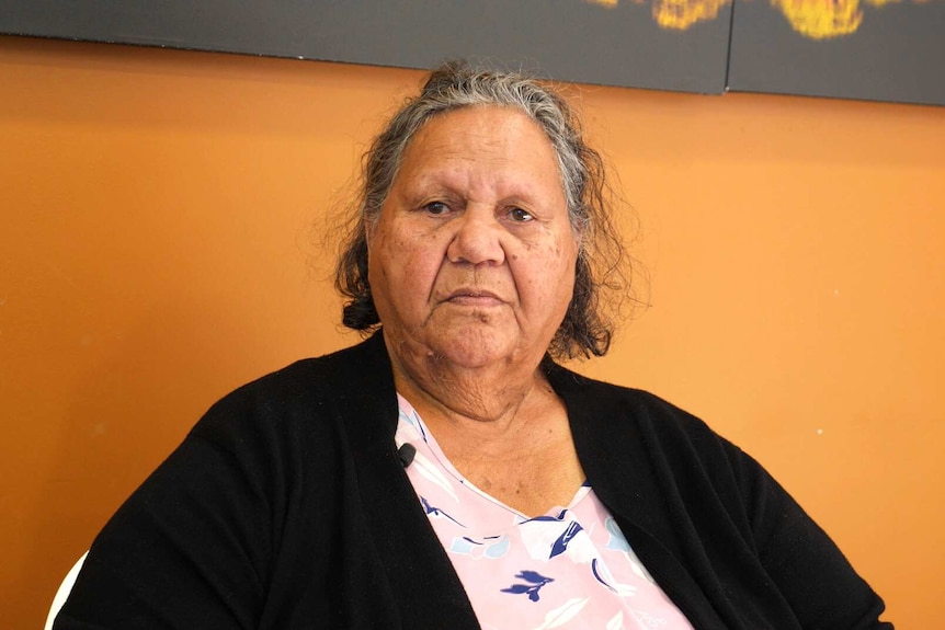 An older lady sits in a chair and looks at the camera with a solemn expression.