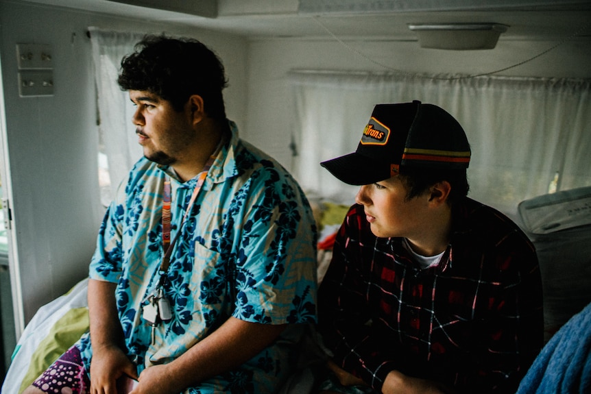 Two teenage boys sit on the bed of a caravan looking out its door, with neutral expressions.