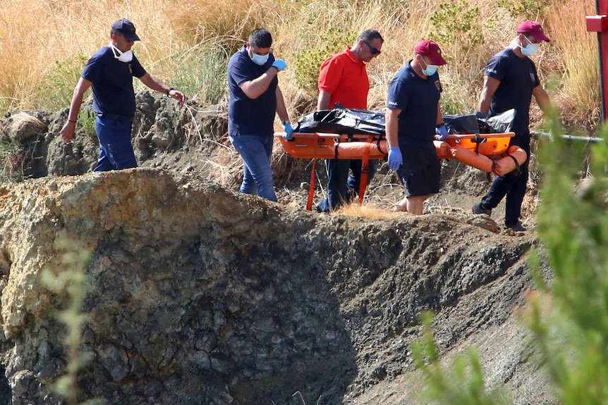 A group of men wearing face masks and gloves carry a black body bag on a plastic stretcher on the rocky edge of a lake.