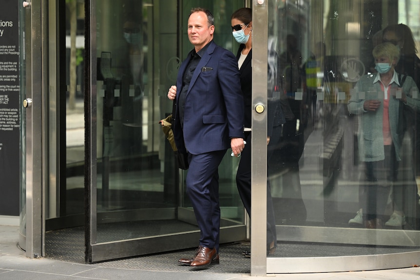 A man wearing a dark blue suit exits a building through glass doors.