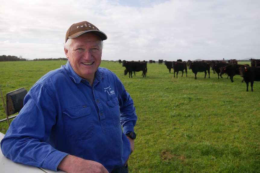 A smiling man in a cap and work shirt stands in a paddock in front of some cattle.