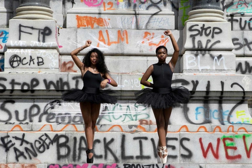Two African American ballerinas standing on the base of a statue covered in graffiti