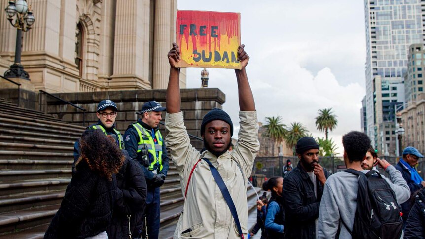 A man in a beige jacket holds a 'Free Sudan' sign high above his head in front of a crowd beside the Victorian Parliament.