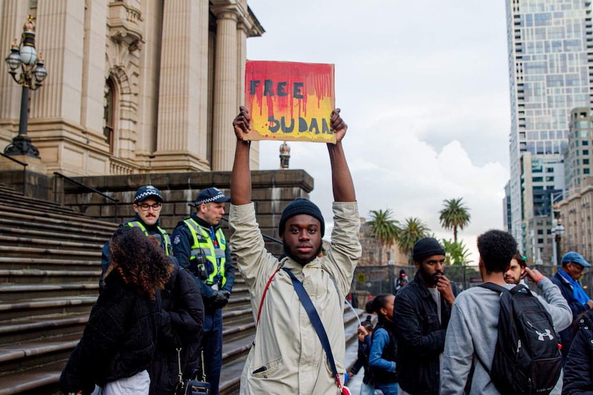 A man in a beige jacket holds a 'Free Sudan' sign high above his head in front of a crowd beside the Victorian Parliament.