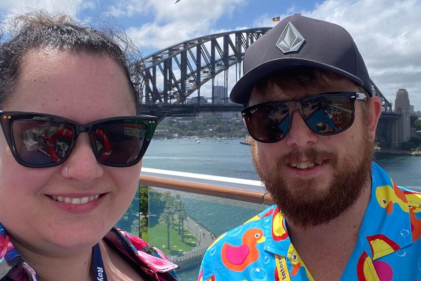 A woman and man wearing sunglasses on a cruise ship in front of the Sydney harbour bridge, they are holding cocktails