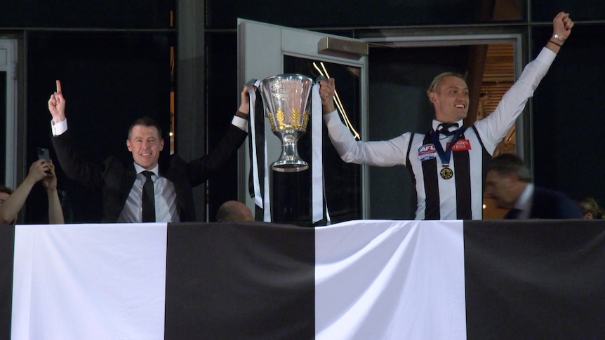 Darcy Moore wears his Collingwood guernsey and medal and holds up the cup with his fist in the air.