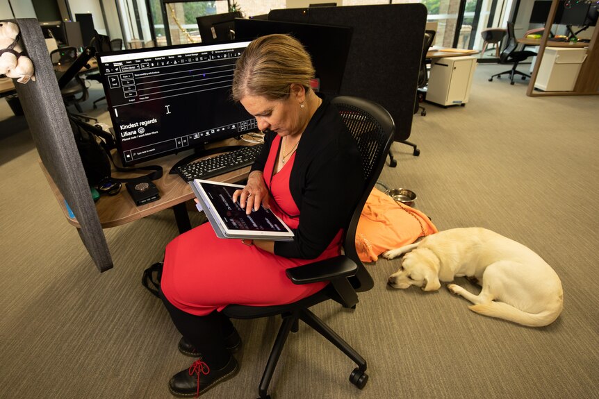 A woman in a red dress sits at a desk using a large iPhone, her guide dog sitting at her feet.
