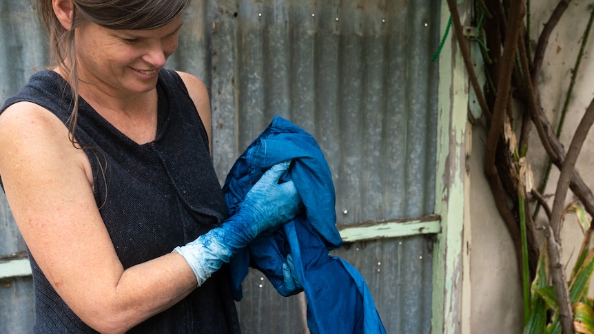 A woman stands in front of a corrugated iron fence while holding blue fabrics and wearing gloves with blue dye on them. 