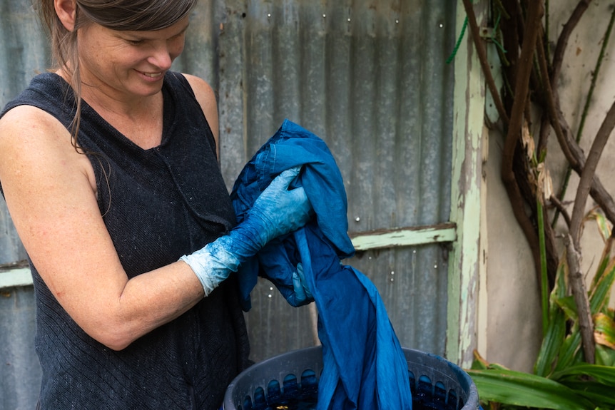 A woman stands in front of a corrugated iron fence while holding blue fabrics and wearing gloves with blue dye on them. 