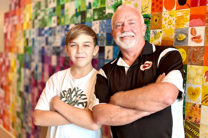 A boy and his father with their arms crossed, smiling, in front of a colourful wall.