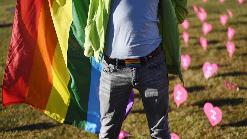 A man stands holding a rainbow flag in front of pink paper hearts decorating a grassy space.