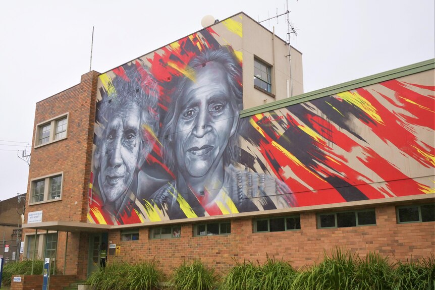 A mural of two Aboriginal women painted in black and white framed by red, black and yellow shading on a brick building