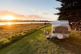A family sits near their caravan, watching the sun set over a river.