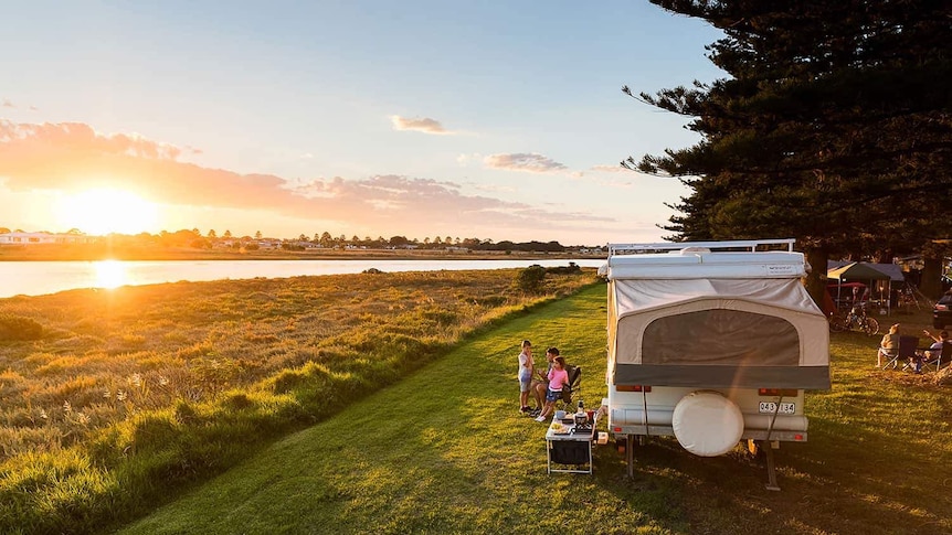 A family sits near their caravan, watching the sun set over a river.