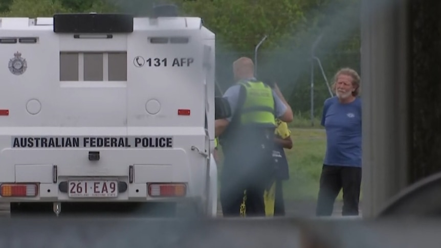 A bearded man with long hair stands near an Australian Federal Police vehicle.