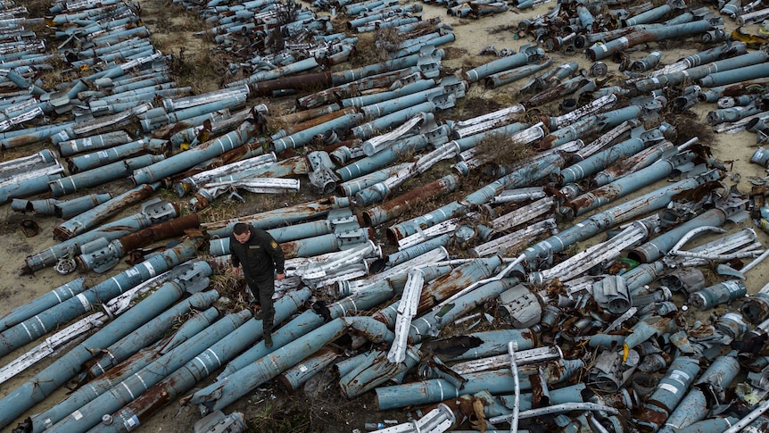 A man in Ukrainian army fatigues walks over a mass of blue metal cylinders lying in a scrapyard.