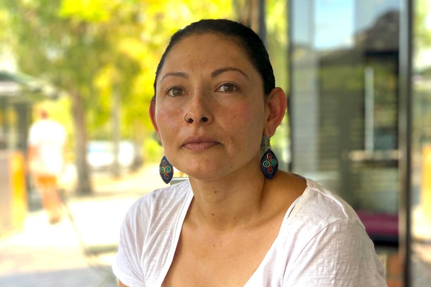 A woman with dark hair and a white shirts sits at a cafe, staring directly into the camera.