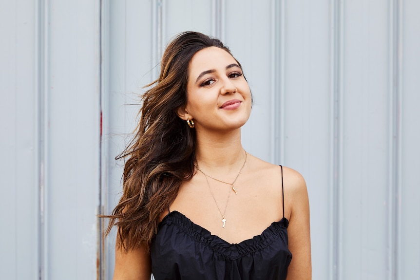 An Italian Australian woman smiles into the camera. She has long brown windswept hair, and her chin raised. 