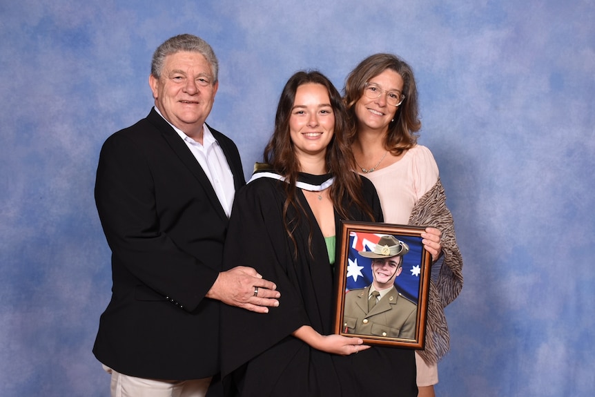 A middle-aged couple with their daughter in graduation robes and holding a framed photograph of a young man in army uniform