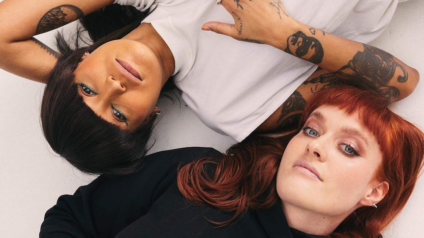 Two women lying on the floor. One has tan skin and dark hair with a fringe, the other has red hair and wears a black turtleneck.