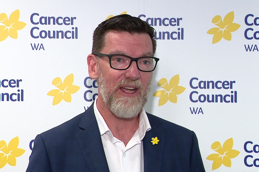 A man with a blue blazer and white collared shirt wears glasses in front of a Cancer Council WA backdrop