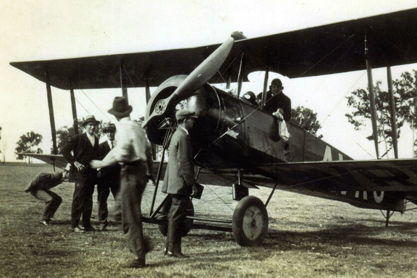 Black and white photo taken in the 1920s of a bi plane in a field with a passenger holding a white mail bag.
