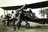 Black and white photo taken in the 1920s of a bi plane in a field with a passenger holding a white mail bag.