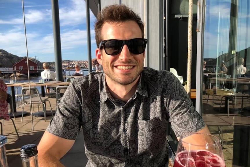 Simon Keane, wearing sunglasses, sits at an outside table at a restaurant on a sunny day with a drink in front of him.