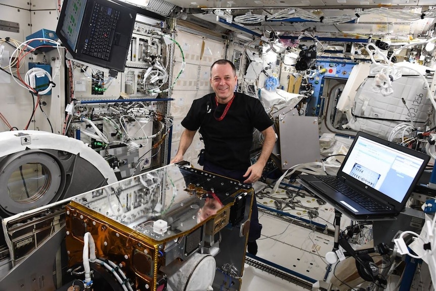 An astronaut smiles with a large metallic box while standing in a room filled with intricate machinery.