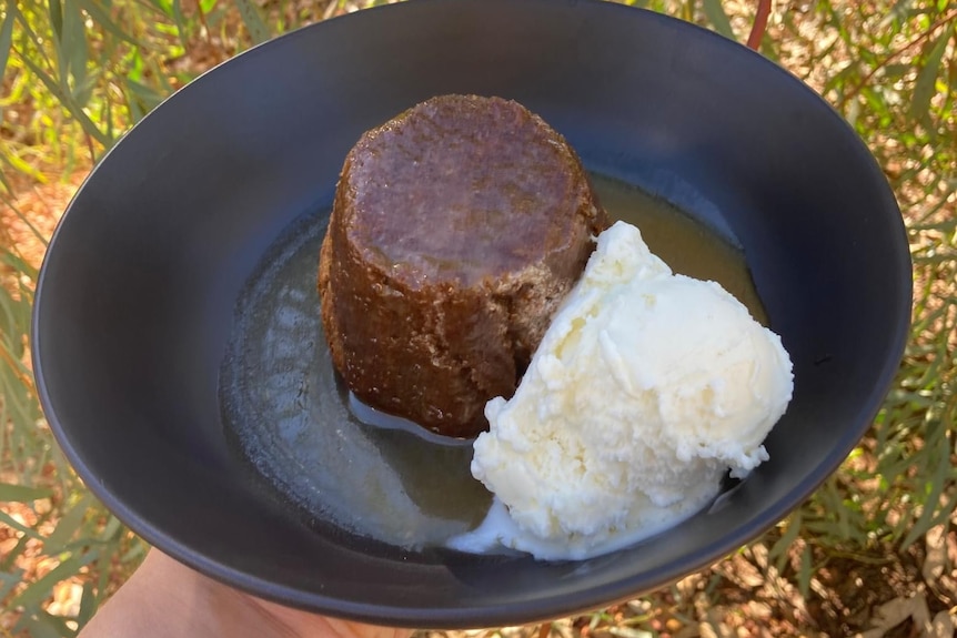 A black bowl with an upturned sticky date pudding (in the shape of a Dariole mold) and a scoop of vanilla ice cream