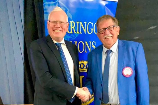 Two men standing together shaking hands in front of a Lions sign.