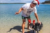 A man in a Christmas hat with his trusty dog, wearing antlers, antlers smiles at the beach.