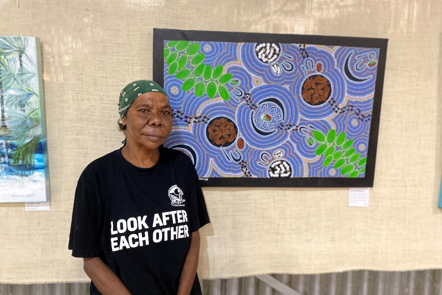 an aboriginal woman wearing a black shirt standing next to a purple and green painting