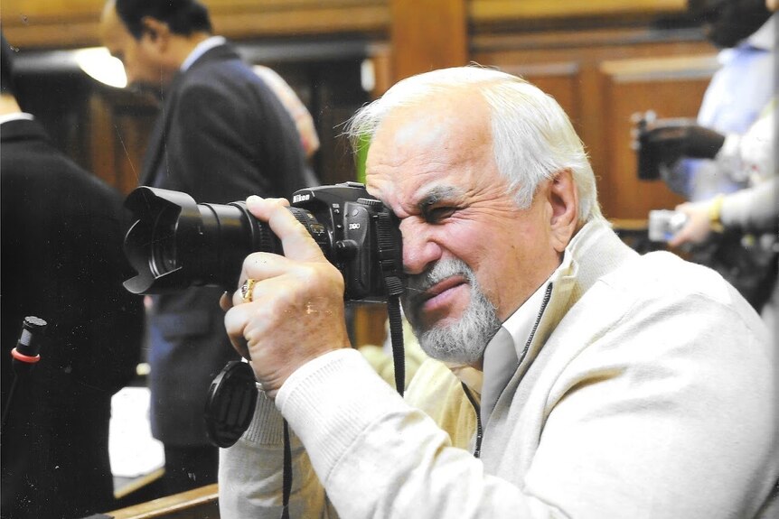 Keith Schafferius wears white and photographs someone with a big black camera in a courtroom