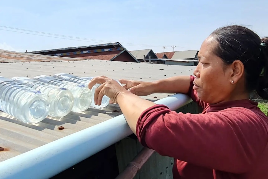 A serious Asian woman wearing a maroon shirt places water bottles on the roof of a house, hair tied back.