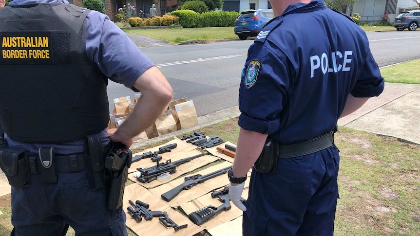 An ABF officer and NSW Police officer stand in front of several weapons on the front lawn of a house in Charlestown.