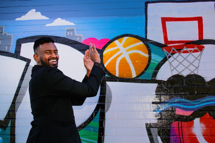 A man stands next to a mural pretending to score a basketball goal.