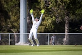 A man wearing cricket whites and bright green wicket keepers' gloves with his hands in the air and mouth open